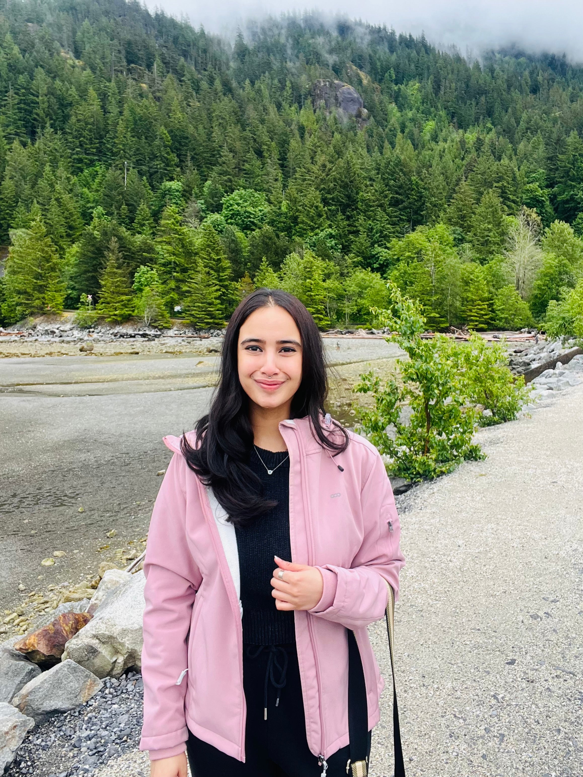 Talk for 5 founder Alisha Jain in a pink jacket standing in a forested area with trees and rocks in the background
