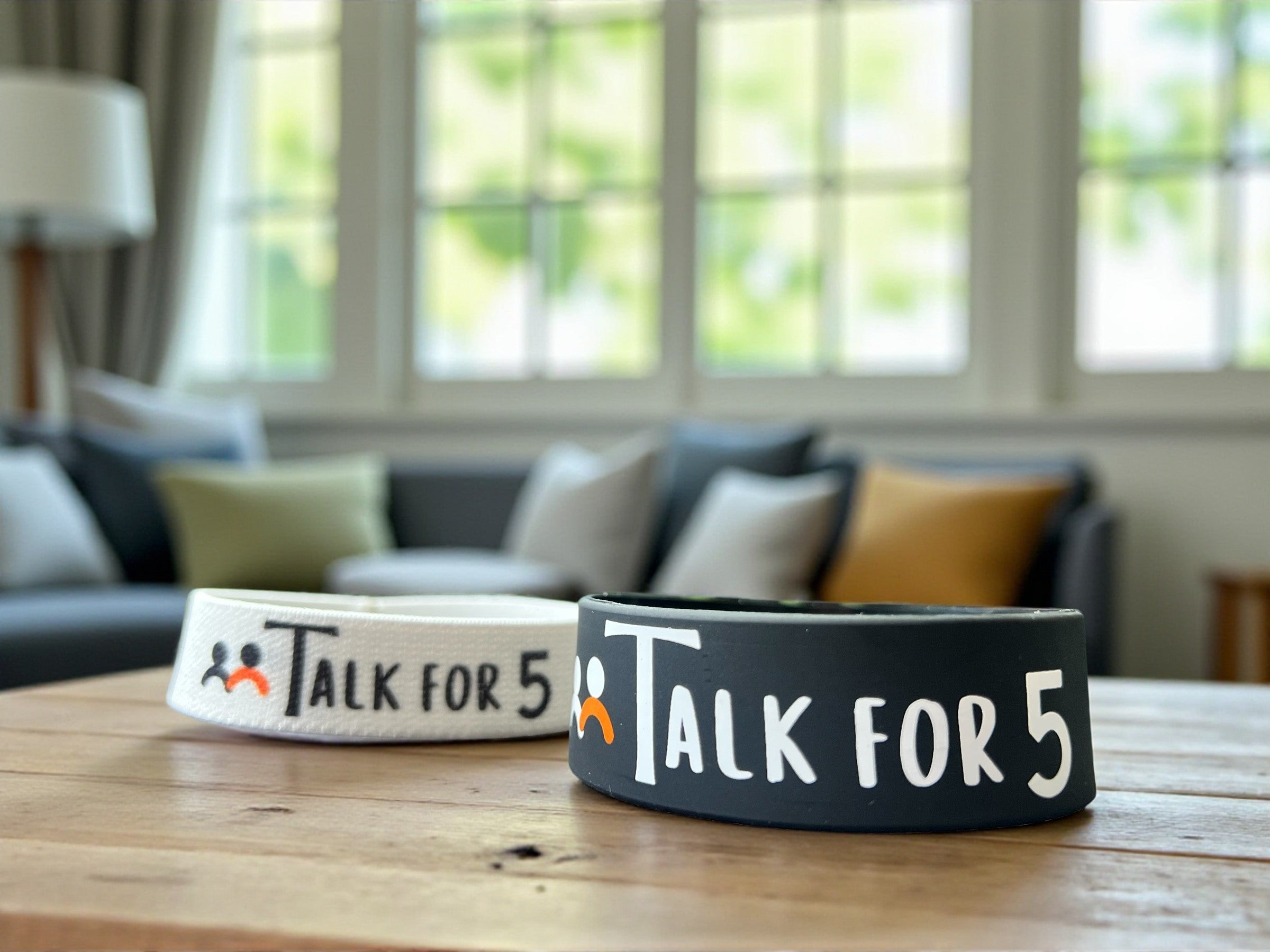 Two 'Talk for 5' conversation starter wristbands on a wooden table with a blurred living room background