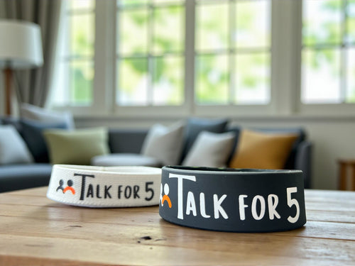 Two 'Talk for 5' conversation starter wristbands on a wooden table with a blurred living room background