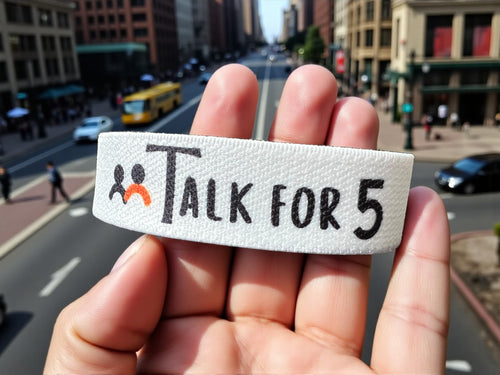 Hand holding a wristband with 'Talk For 5' text against an urban street background where people are walking and driving