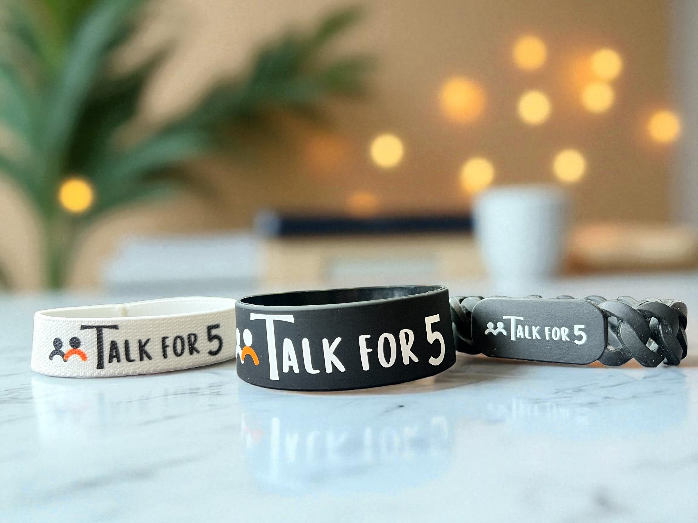 Three Talk for 5 wristbands displayed on a marble surface with a plant and lightings in the background.