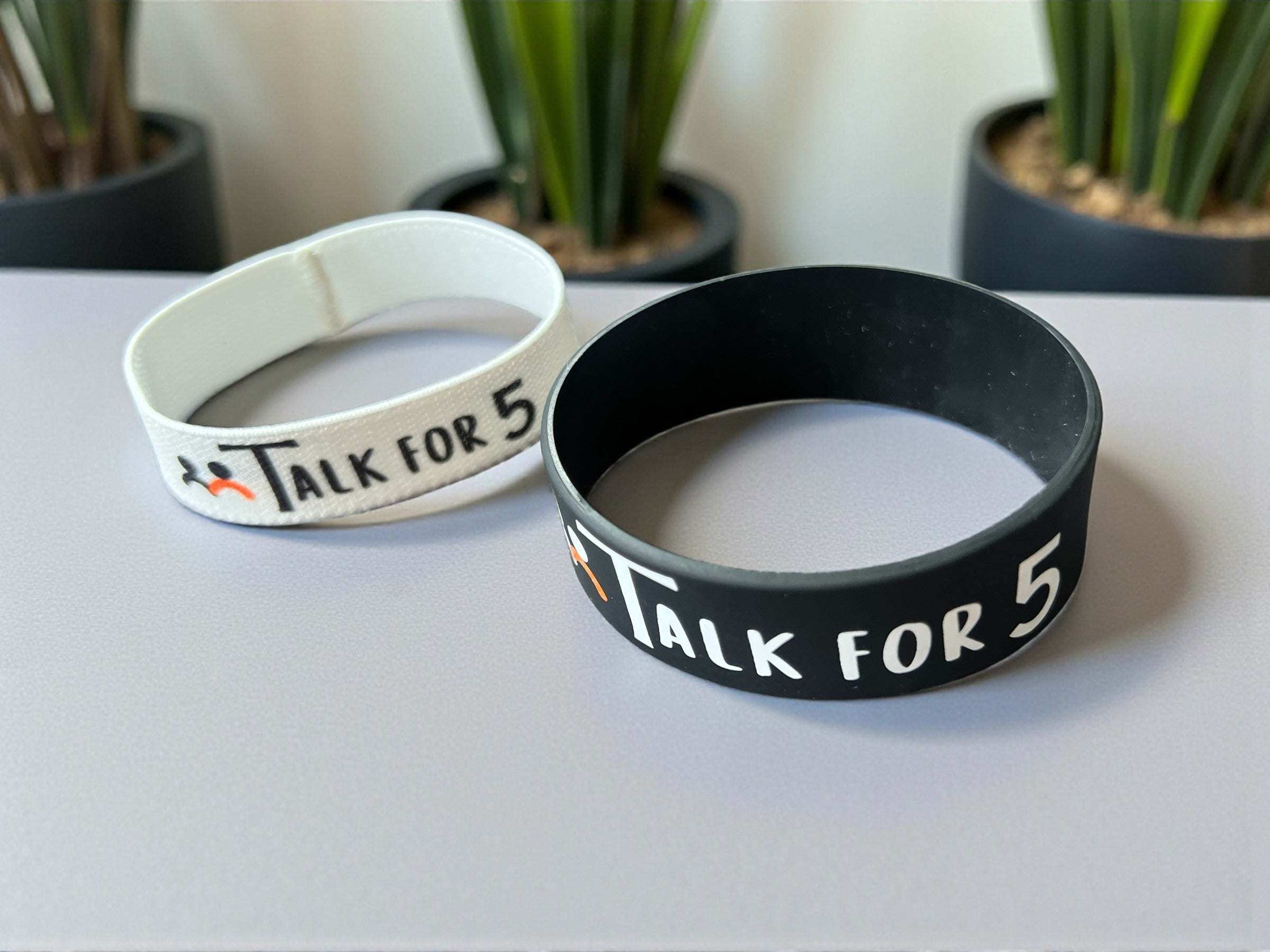 Two mental health awareness wristbands with 'Talk For 5' text on a light surface with plants in the background.