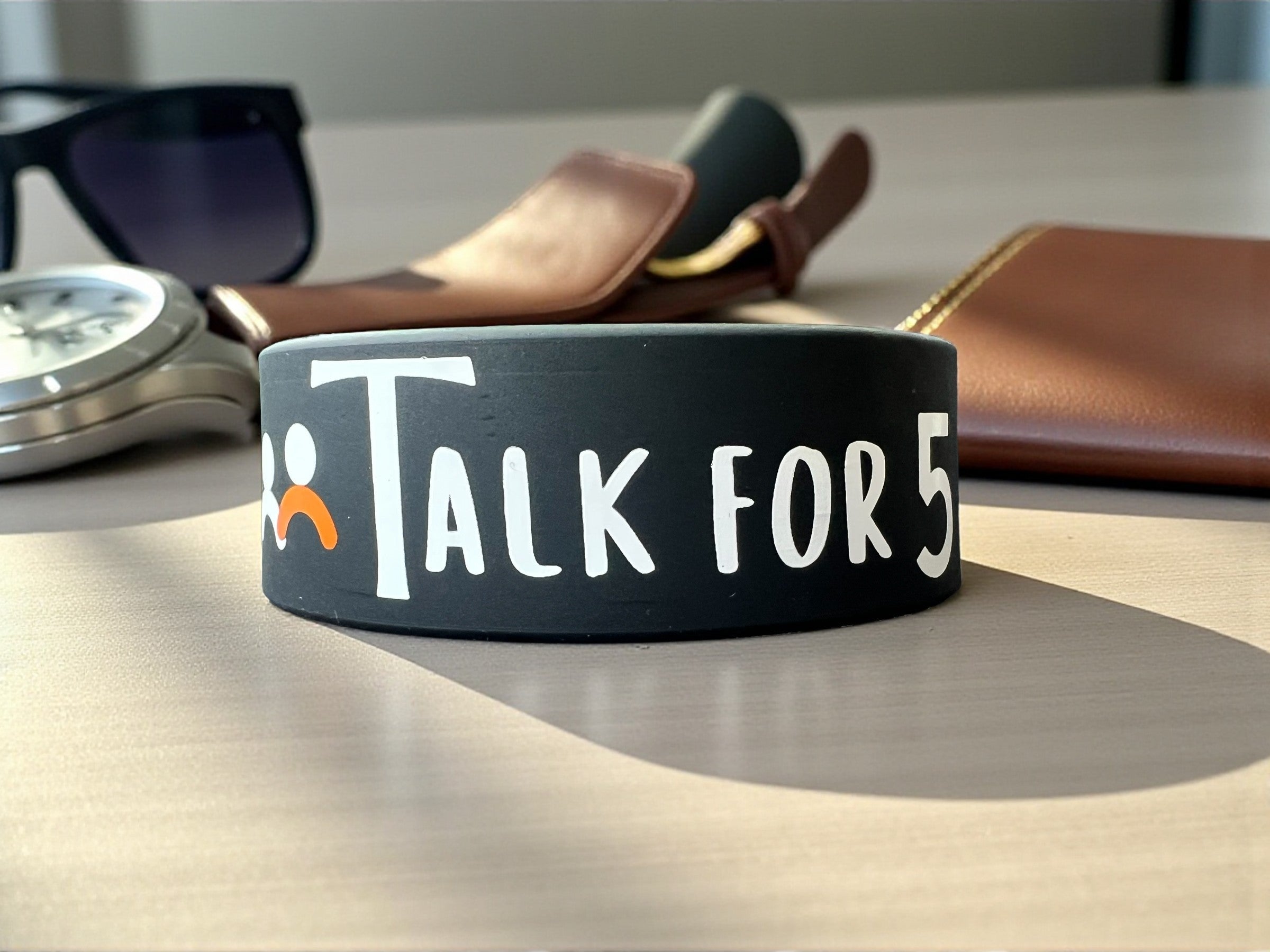 Black wristband with 'Talk For 5' text on a table with sunglasses and wallet in the background