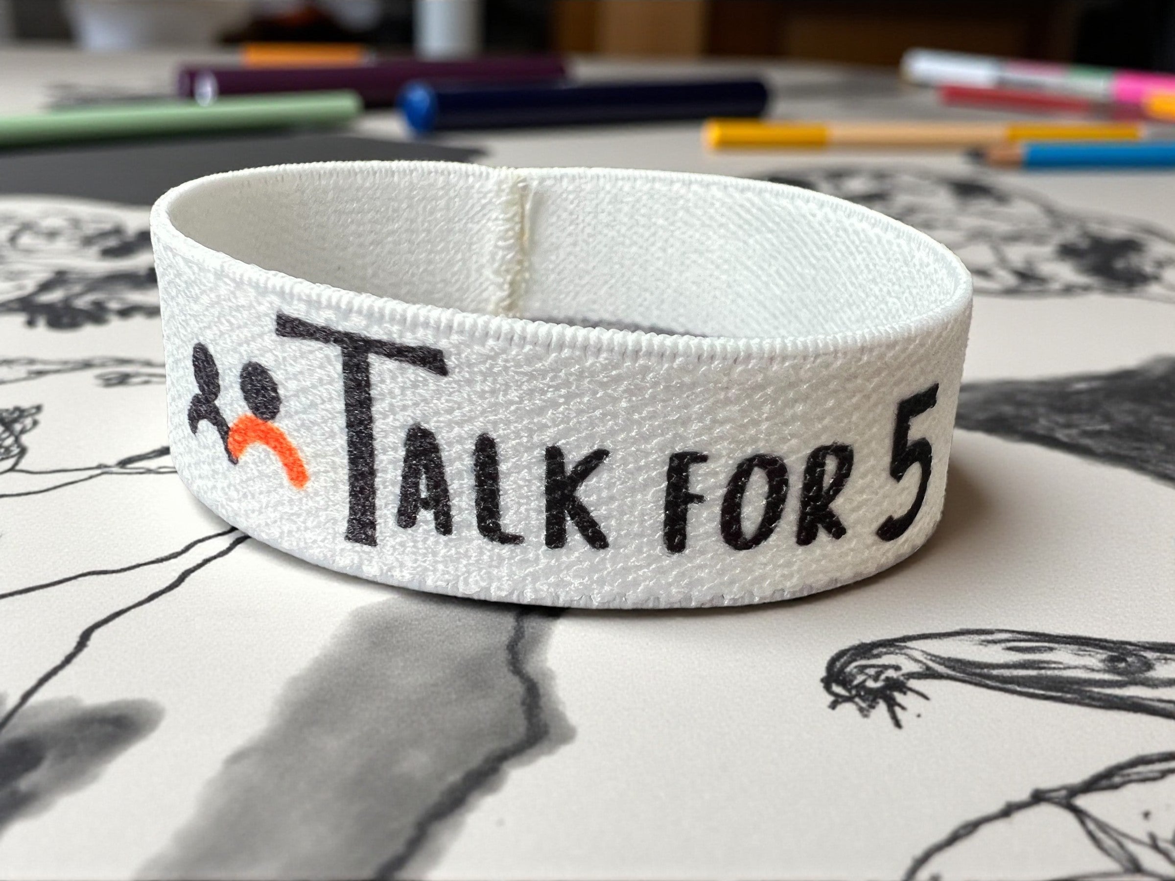 White light-weight hand accessory style Talk for 5 wristband to support mental wellbeing, placed on an artistic table